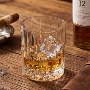 May include: A rocks glass with ice and amber liquid, a bottle of The Macallan 12 Year Old Scotch, a cigar in an ashtray, and a leather-bound notebook with a pen on a wooden surface.