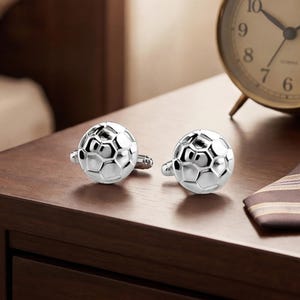 May include: A pair of silver cufflinks with a soccer ball design, next to a brown and tan striped tie and a gold-colored alarm clock. The items are arranged on a dark brown wooden nightstand.