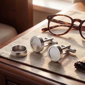 Puede incluir: Gemelos plateados con diseño circular, un anillo plateado y gafas de carey sobre una mesita de noche de madera. También hay un paño estampado. La escena está iluminada por luz natural.