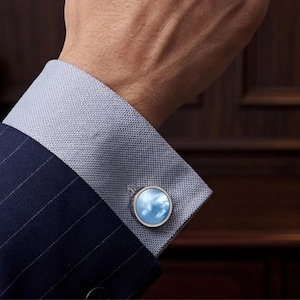 May include: A close-up of a blue pinstripe suit with a light blue dress shirt and a silver cufflink with a blue sky design. The cufflink is round and has a silver border. The suit is dark blue.