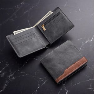 Puede incluir: Dos carteras grises, una abierta y otra cerrada, sobre una superficie oscura. La cartera abierta revela un compartimento para efectivo con cremallera y tirador dorado. La cartera cerrada tiene un acento marrón y costuras blancas.