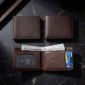 May include: Three brown leather wallets are displayed on a dark surface. One wallet is open, revealing cash and credit cards. The open wallet has a clear window for a card with the text "BLACK UNIQUE" and a card number. The other two wallets are closed.
