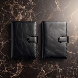 Könnte beinhalten: Zwei schwarze Lederjournale mit Schnappverschluss werden auf einer dunklen, marmorierten Oberfläche gezeigt. Die Journale sind rechteckig, mit abgerundeten Ecken und einem genähten Rand. Der Schnappverschluss ist gebogen.