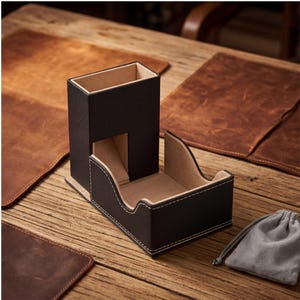 May include: A black and beige card holder and dice tower set. The set includes a rectangular tower with a cutout and a curved tray, both with white stitching. A small gray bag is also included. The set is on a wooden surface.