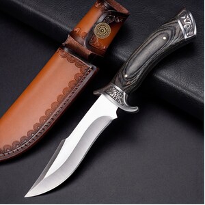 May include: A hunting knife with a polished steel blade and a dark wood handle. The knife features a decorative metal bolster and is accompanied by a brown leather sheath with embossed detailing. The sheath has a decorative pattern.