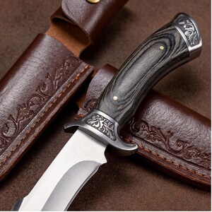 May include: A hunting knife with a silver blade and a dark wood handle. The handle has a decorative metal bolster and end cap. The knife is set in a brown leather sheath with embossed floral designs.