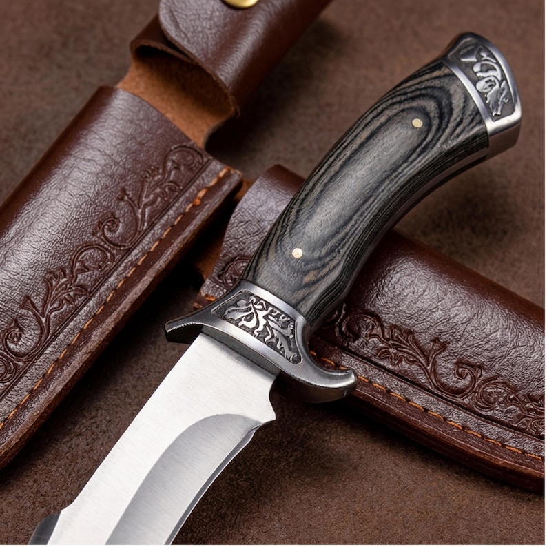 May include: A hunting knife with a silver blade and a dark wood handle. The handle has a decorative metal bolster and end cap. The knife is set in a brown leather sheath with embossed floral designs.