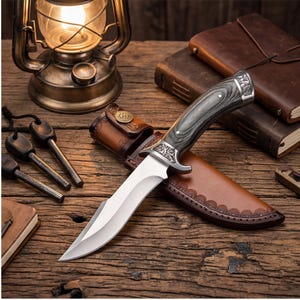 May include: A hunting knife with a silver blade and a dark wood handle rests in a brown leather sheath. The knife is on a wooden surface with a vintage lantern, fire starters, and leather-bound books.