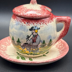 May include: A vintage ceramic tea cup and saucer set. The cup features a hand-painted scene of a man smoking a pipe, with a red and white speckled lid. The saucer has a matching red and white speckled rim and green leaf accents.