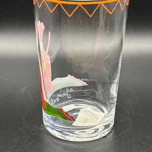May include: A clear glass tumbler with an orange zig-zag design around the rim. The glass features a cartoon illustration of a pink panther with a green and red design. The glass has a signature on the bottom.