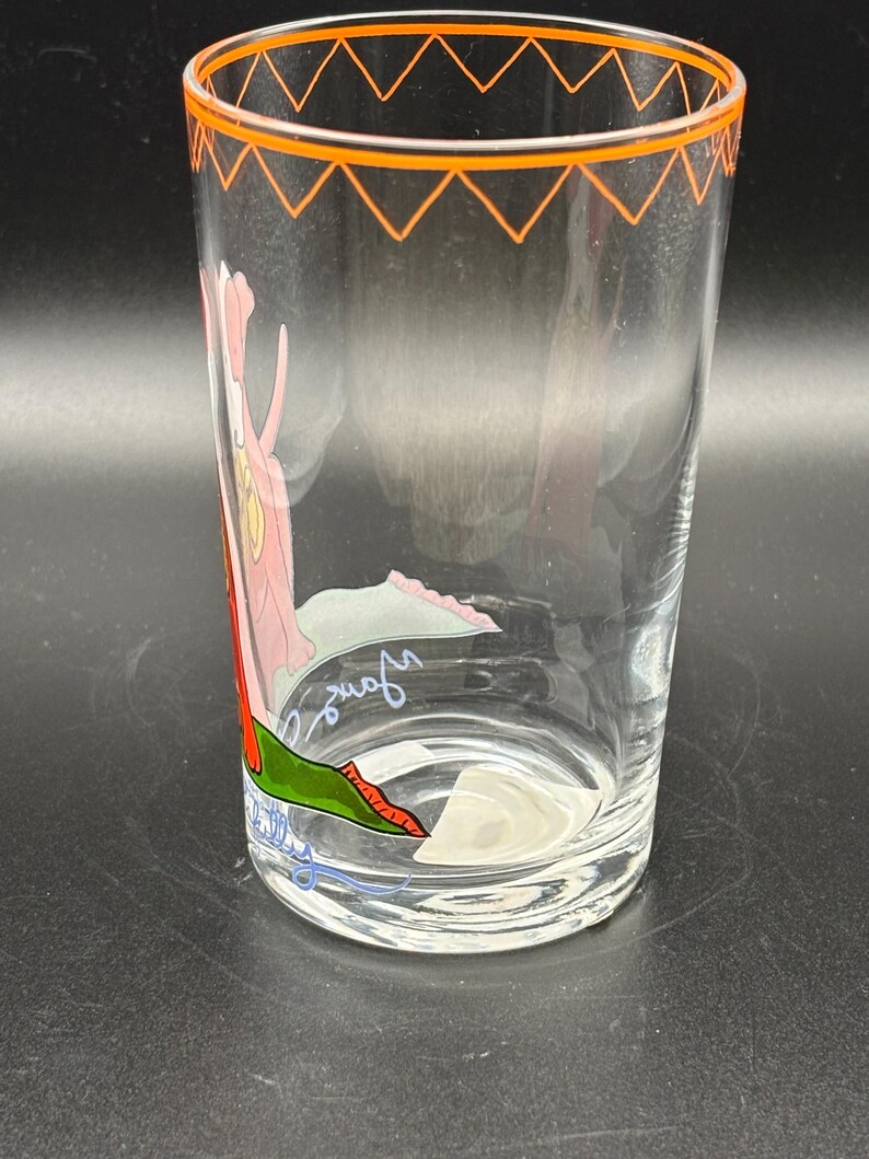 May include: A clear glass tumbler with an orange zig-zag design around the rim. The glass features a cartoon illustration of a pink panther with a green and red design. The glass has a signature on the bottom.