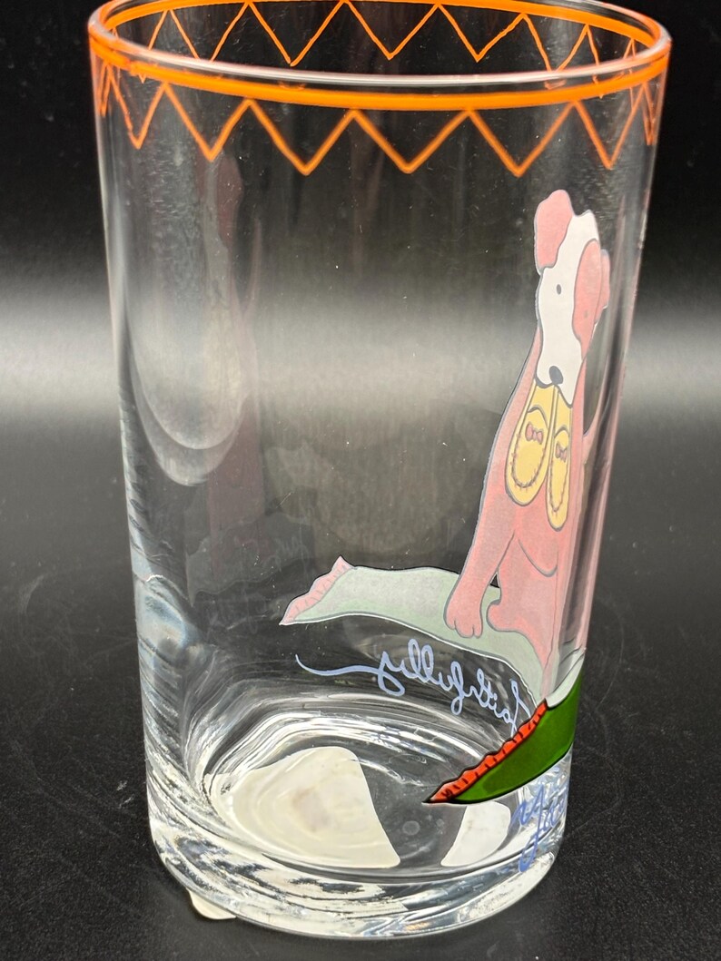 May include: Clear glass tumbler with an orange zig-zag design around the rim. The glass features a cartoon dog illustration in pink, white, and yellow. The dog is sitting on a green mat. The word "jellyfish" is written on the glass.