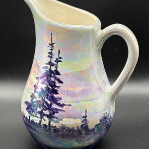 May include: Iridescent ceramic pitcher with a landscape painting. The pitcher features a handle and spout. The landscape painting depicts purple trees and mountains with a colorful sky. The artist's signature is visible on the bottom of the pitcher.
