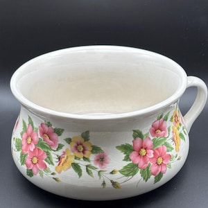 May include: A cream-colored ceramic chamber pot featuring a floral pattern of pink and yellow flowers with green leaves around the exterior. The vessel has a wide, rounded bowl shape, a smooth interior, and a single handle on the right side.
