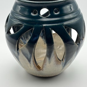 May include: A round ceramic candle holder featuring a glossy dark teal glaze on the upper portion that transitions into a light beige base. The vessel has a series of petal-shaped cutouts around the center and small circular holes near the rim.