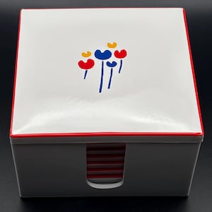 May include: A square white box with a red border and a graphic of stylized blue, red, and yellow flowers on the lid. The front of the box features a U-shaped cutout revealing a stack of red-edged square coasters stored inside the container.