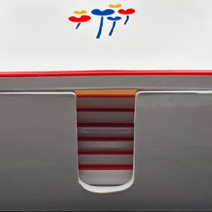 May include: A white square coaster holder with a red trim and a cutout front opening. The top surface features a graphic of small red, blue, and yellow heart-shaped balloons. Inside the holder, a stack of white coasters with red edges is visible.