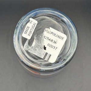 May include: Clear glass drinking glass with a blue rim. The bottom of the glass has a white sticker that says "MADE IN PORTUGAL" and another sticker with the word "ANTHROPOLOGIE" and a barcode.