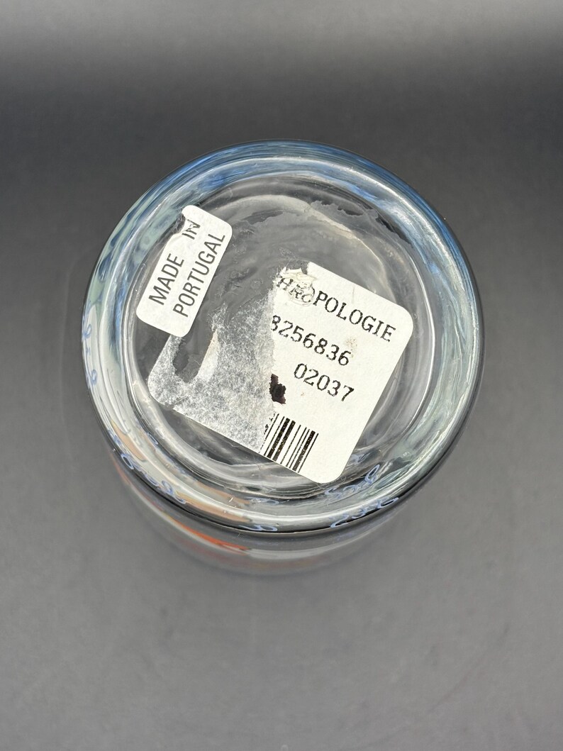 May include: Clear glass drinking glass with a blue rim. The bottom of the glass has a white sticker that says "MADE IN PORTUGAL" and another sticker with the word "ANTHROPOLOGIE" and a barcode.