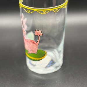 May include: Clear glass tumbler with a yellow decorative rim. The glass features a pink and red illustration of a flamingo-like creature on a green base. The glass is cylindrical and appears to be hand-painted.