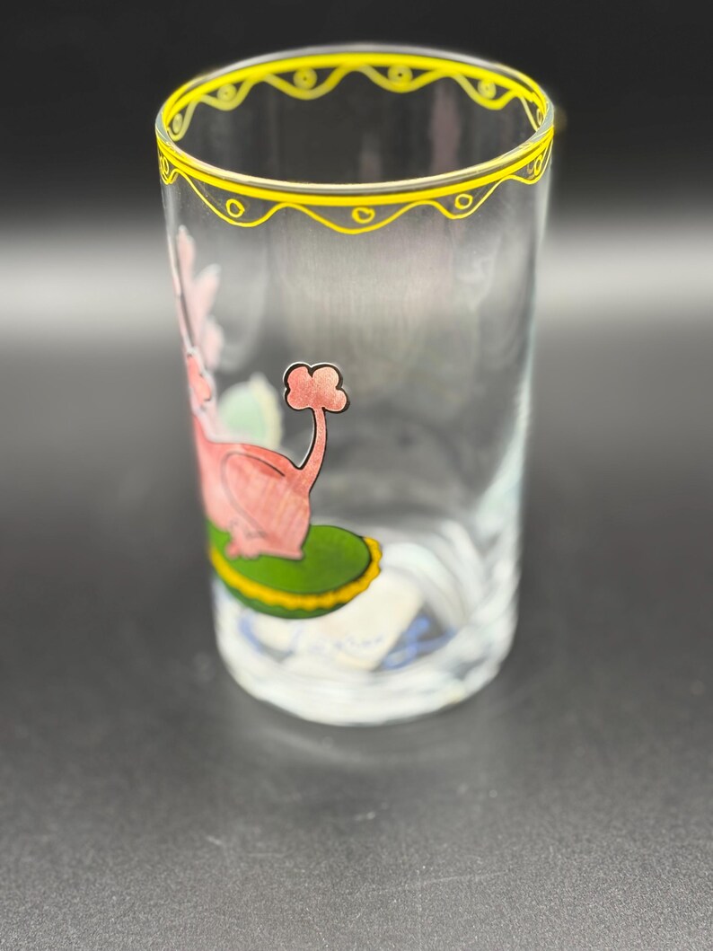 May include: Clear glass tumbler with a yellow decorative rim. The glass features a pink and red illustration of a flamingo-like creature on a green base. The glass is cylindrical and appears to be hand-painted.