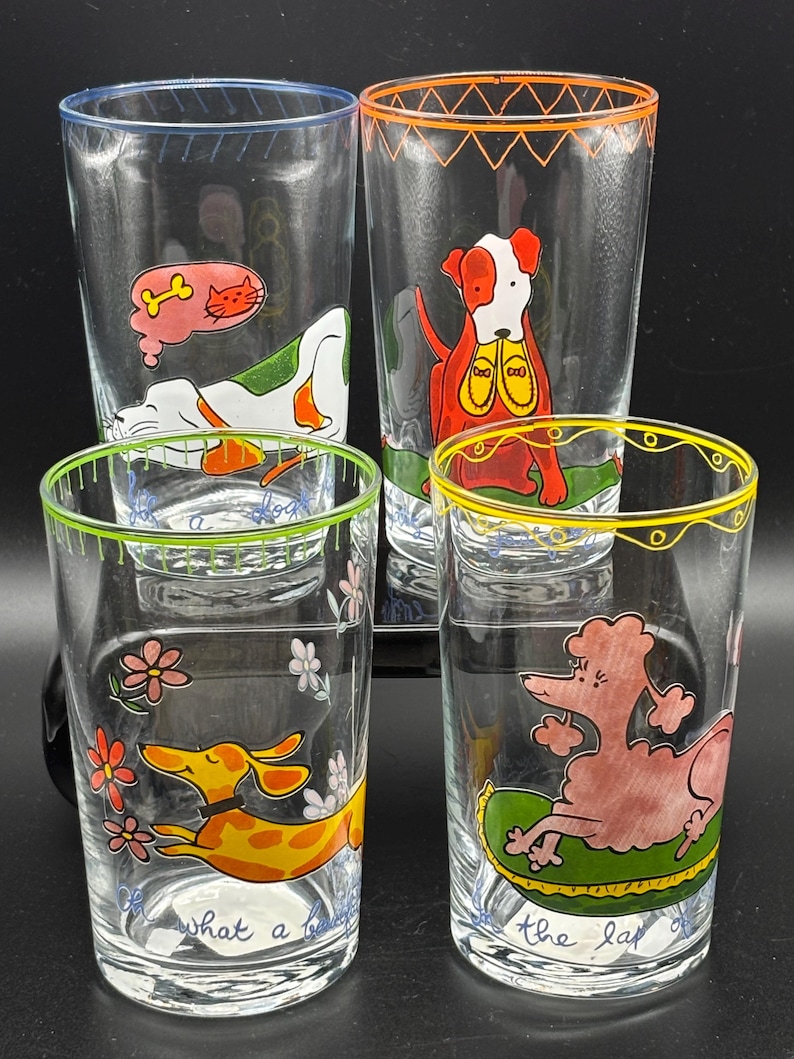 May include: Set of four clear drinking glasses with colorful dog illustrations. Each glass features a different dog design, with accents of blue, orange, green, and yellow rims. One glass has the text "what a" and another has "the lap of".