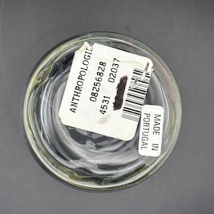 May include: Clear glass container with a white label that reads "ANTHROPOLOGIE" and includes a barcode. The label also states "MADE IN PORTUGAL". The container is on a dark gray surface.