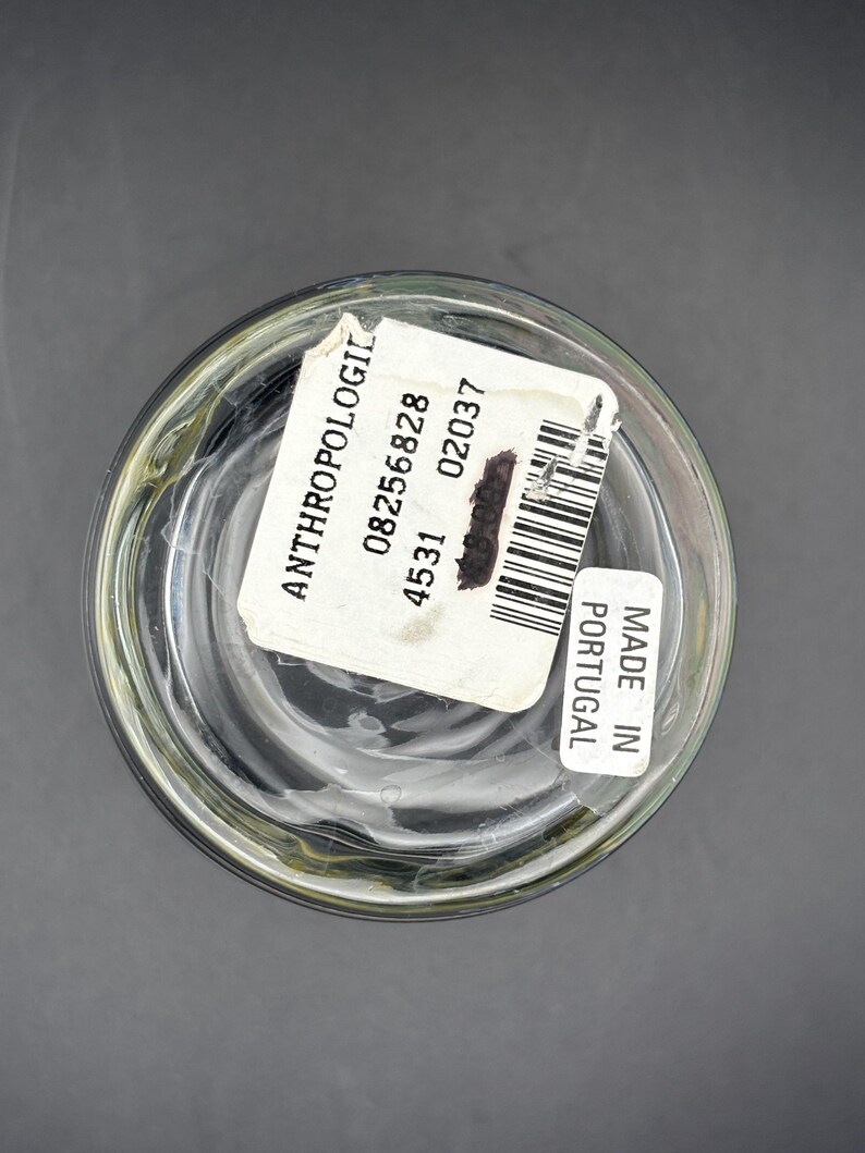 May include: Clear glass container with a white label that reads "ANTHROPOLOGIE" and includes a barcode. The label also states "MADE IN PORTUGAL". The container is on a dark gray surface.