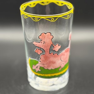 May include: A clear glass with a yellow rim and a pink poodle illustration. The poodle is lying on a green and yellow surface. The glass has a white base with a cloud-like design.