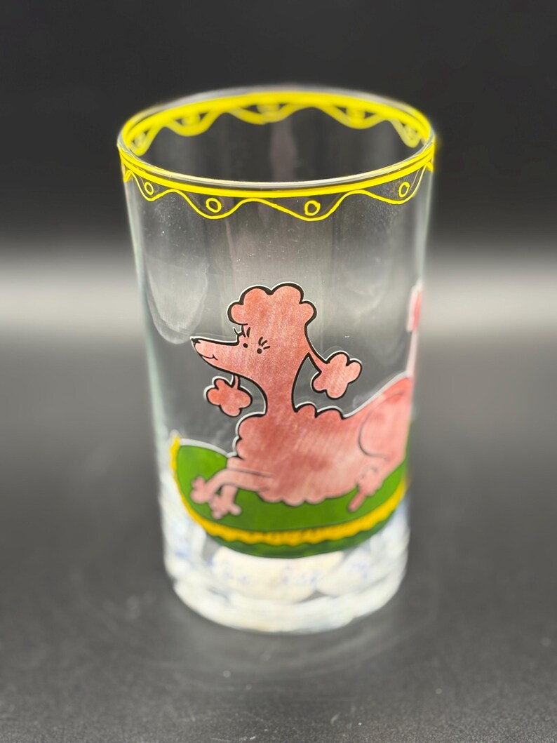 May include: A clear glass with a yellow rim and a pink poodle illustration. The poodle is lying on a green and yellow surface. The glass has a white base with a cloud-like design.