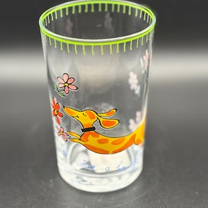 May include: Clear glass tumbler with a green rim and a whimsical design. The tumbler features a cartoon dachshund in orange with yellow spots, a black collar, and pink and orange flowers. The words "share a smile" are at the bottom.