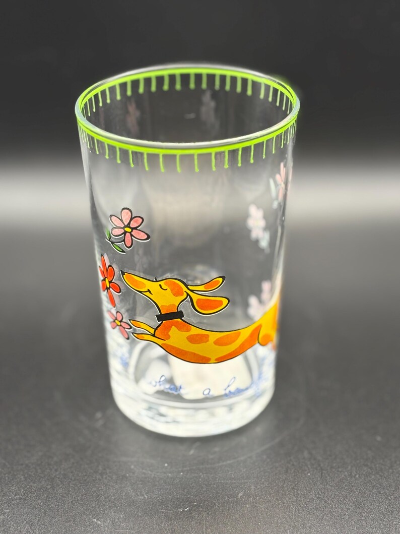 May include: Clear glass tumbler with a green rim and a whimsical design. The tumbler features a cartoon dachshund in orange with yellow spots, a black collar, and pink and orange flowers. The words "share a smile" are at the bottom.