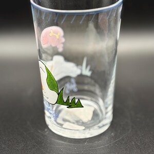 May include: A clear glass tumbler with a blue rim and cartoon illustrations. The design includes a pink pig with a bone, a white cloud, and green grass. The glass is cylindrical and appears to be empty.