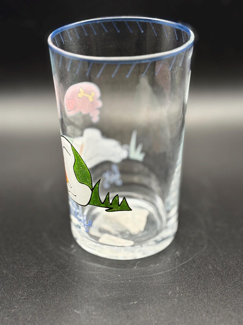 May include: A clear glass tumbler with a blue rim and cartoon illustrations. The design includes a pink pig with a bone, a white cloud, and green grass. The glass is cylindrical and appears to be empty.