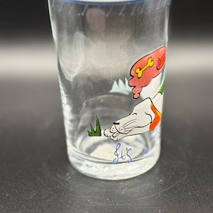 May include: Clear glass tumbler with a blue rim and a cartoon dog design. The dog is white with orange and red accents, lying down with a bone and cat graphic. The glass has a handwritten signature at the bottom.