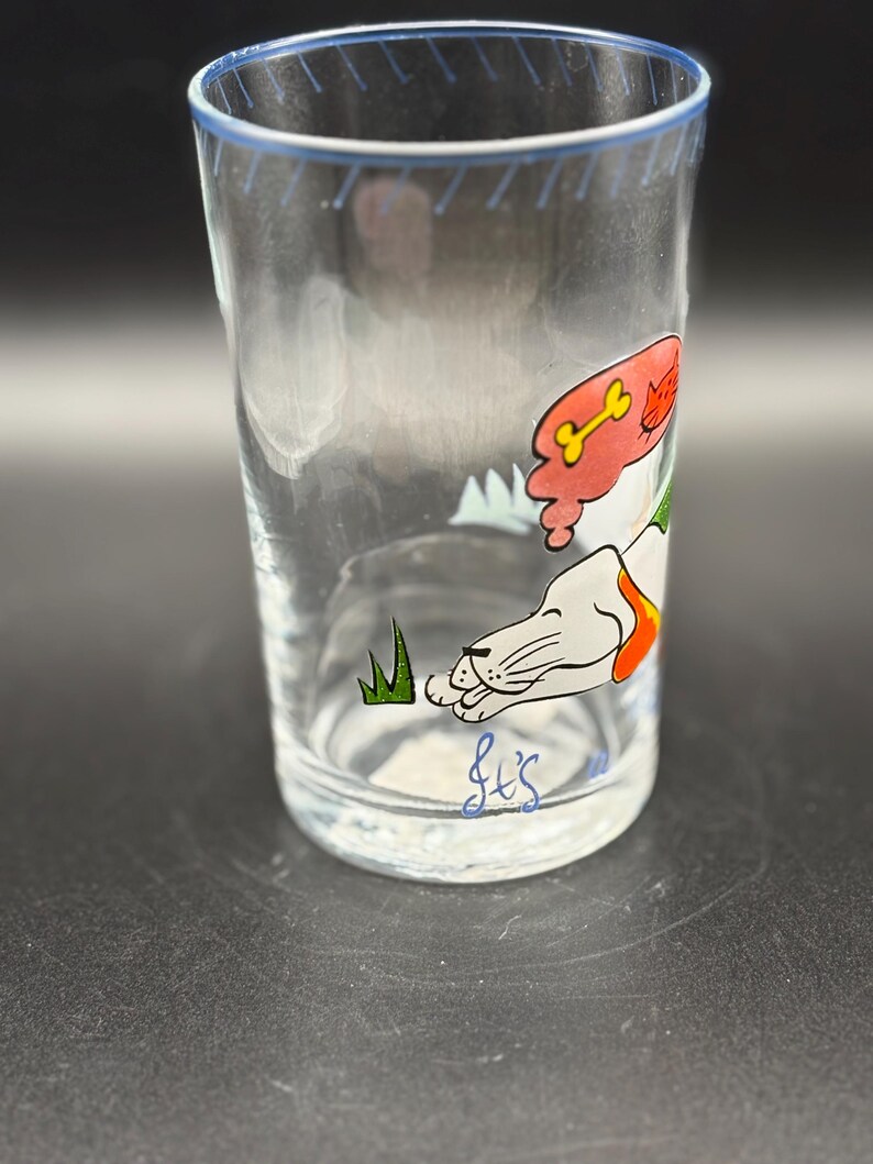 May include: Clear glass tumbler with a blue rim and a cartoon dog design. The dog is white with orange and red accents, lying down with a bone and cat graphic. The glass has a handwritten signature at the bottom.