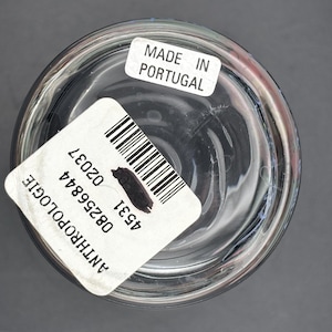 May include: Clear glass container with a rounded base. A white label with a barcode and the text "ANTHROPOLOGIE" is affixed to the side. Another label reads "MADE IN PORTUGAL". The container is set against a gray background.