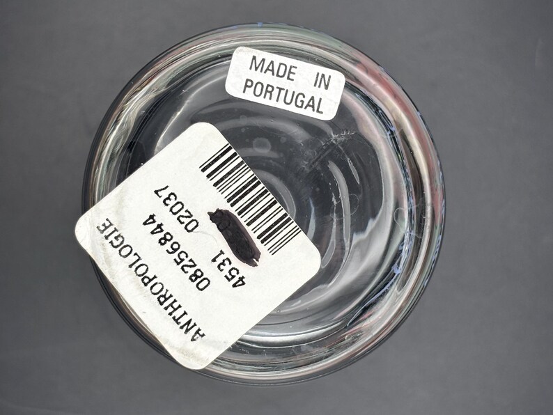 May include: Clear glass container with a rounded base. A white label with a barcode and the text "ANTHROPOLOGIE" is affixed to the side. Another label reads "MADE IN PORTUGAL". The container is set against a gray background.