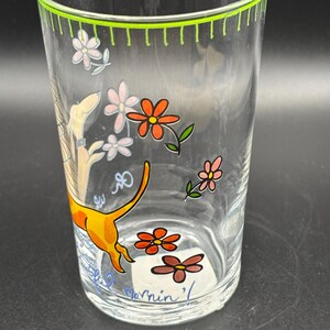 May include: Clear glass tumbler with a green rim and floral and dog illustrations. The tumbler features orange, pink, and brown flowers, and a yellow dog with an orange tail. The text "Mornin'" is visible on the bottom.