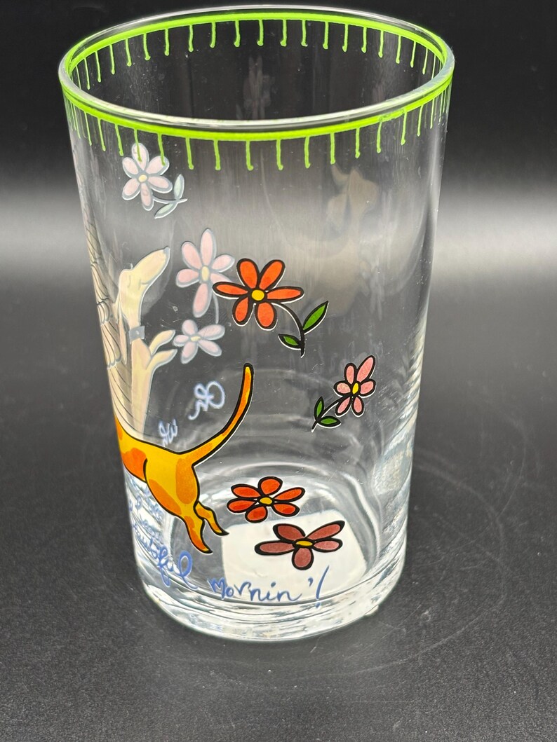 May include: Clear glass tumbler with a green rim and floral and dog illustrations. The tumbler features orange, pink, and brown flowers, and a yellow dog with an orange tail. The text "Mornin'" is visible on the bottom.