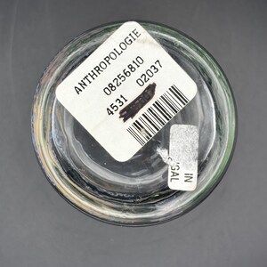 May include: Clear glass Anthropologie drinking glass with a white label. The label includes the brand name, a barcode, and other numbers. The glass is on a dark gray surface.
