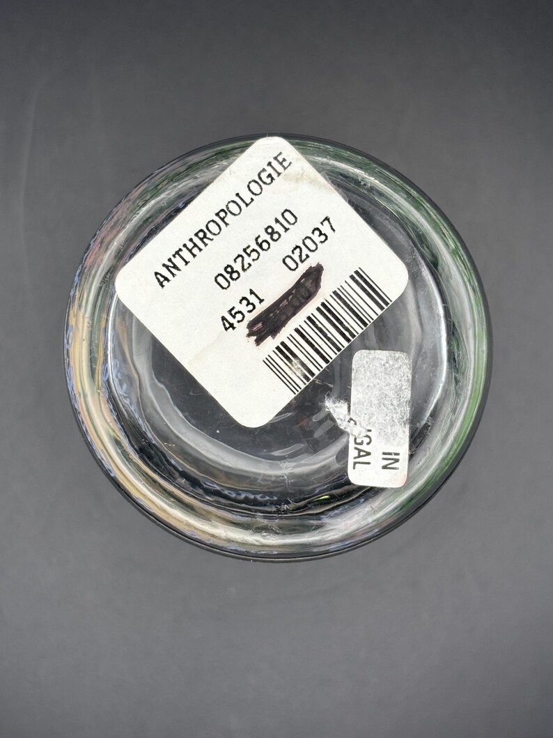 May include: Clear glass Anthropologie drinking glass with a white label. The label includes the brand name, a barcode, and other numbers. The glass is on a dark gray surface.