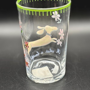 May include: Clear glass tumbler with a green rim and a whimsical design. The tumbler features a cartoon dachshund dog leaping among pink and white flowers. The words "lifeguard a take" are written on the glass.