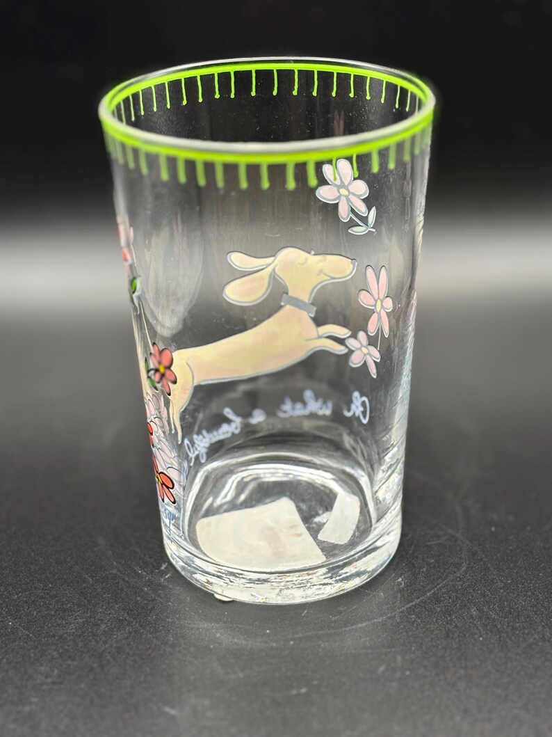 May include: Clear glass tumbler with a green rim and a whimsical design. The tumbler features a cartoon dachshund dog leaping among pink and white flowers. The words "lifeguard a take" are written on the glass.