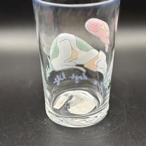 May include: Clear glass tumbler with a blue rim and a whimsical design. The illustration features a sleeping dog with green spots and a pink cat with a bone. The words "yif yab" are written on the glass.