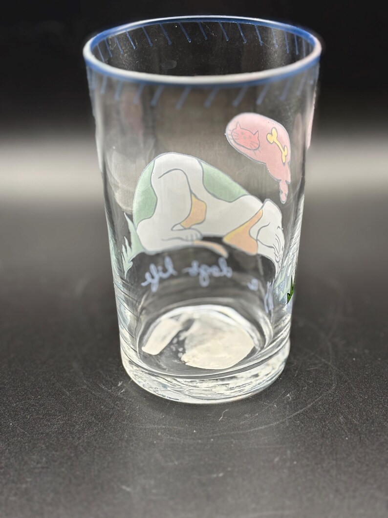 May include: Clear glass tumbler with a blue rim and a whimsical design. The illustration features a sleeping dog with green spots and a pink cat with a bone. The words "yif yab" are written on the glass.