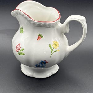 May include: White ceramic pitcher with a scalloped rim and base, adorned with colorful floral and strawberry designs. The pitcher has a curved handle and a red rim, perfect for serving beverages.