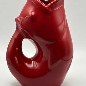 May include: A glossy red ceramic pitcher shaped like a fish with its mouth open at the top. The handle is formed by the curved tail section of the fish, creating a circular opening. The surface is smooth and reflective with a solid red color.