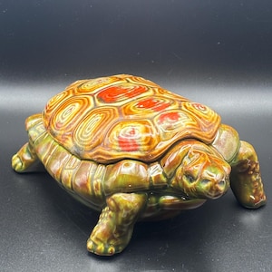 May include: A ceramic turtle-shaped trinket box. The box is glazed in shades of green, brown, and orange, with a detailed shell pattern. The turtle has a detailed face and legs, and the lid fits snugly on the base.