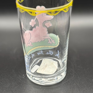 May include: Clear glass tumbler with a pink poodle illustration resting on a green pillow. The rim is decorated with a yellow scalloped design. The glass has text in cursive script near the base.